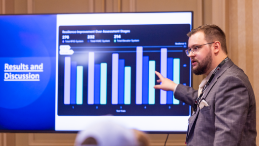 A man stands in front of a screen and presents to a large group about research depicted on the screen in the form of a bar graph.