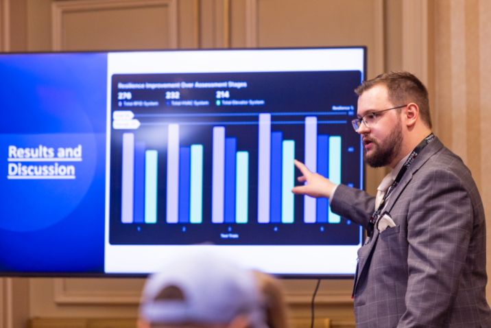 A man stands in front of a screen and presents to a large group about research depicted on the screen in the form of a bar graph.