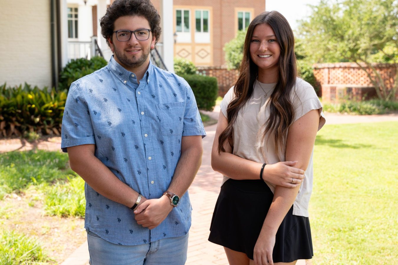 A male student stands outside with his hands folded in front of him while a female student stands to his right with her left hand across her body holding her right arm