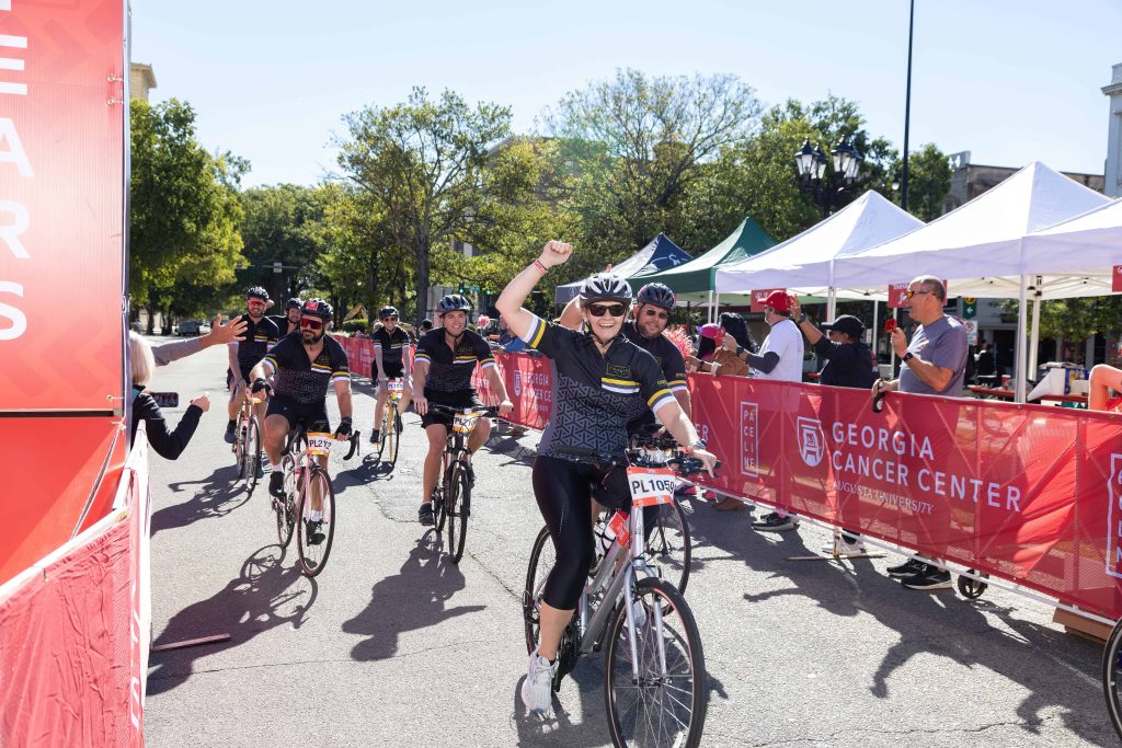 A woman riding on a bicycle through a finish line of a race raising a hand over her head to cheer.