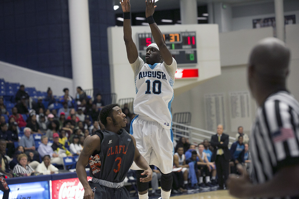 A male college basketball player wearing a white Augusta jersey with the number 10 on it shoots a three-point shot over an opposing player in front of a large crowd.