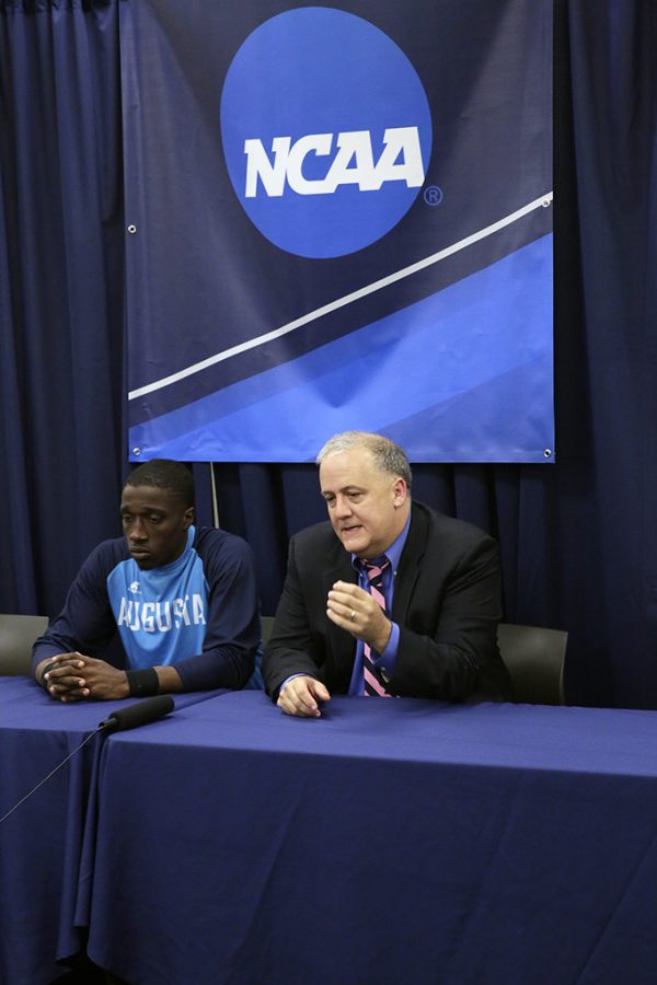 Two men sit at a table during a press conference and speak into a microphone lying on the table. Behind them is a banner with the NCAA logo on it.