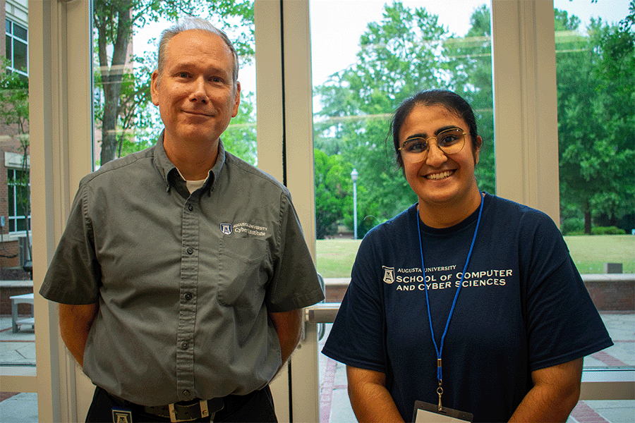 A man in an Augusta University Cyber Institute shirt stands with a woman wearing an Augusta University School of Computer and Cyber Sciences tshirt as they pose for a photo in front of a wall of windows.