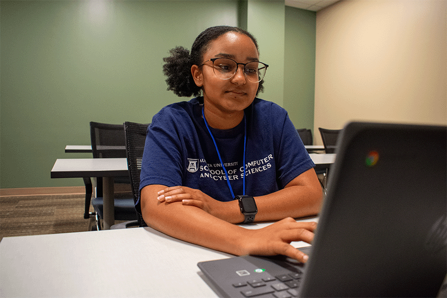 A woman in a navy Augusta University School of Computer and Cyber Sciences tshirt sits at a table in front of a laptop that she's working on.