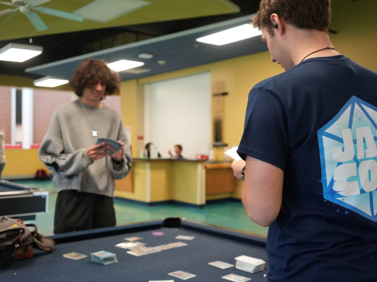 Two male college students play a card game while standing next to a pool table.