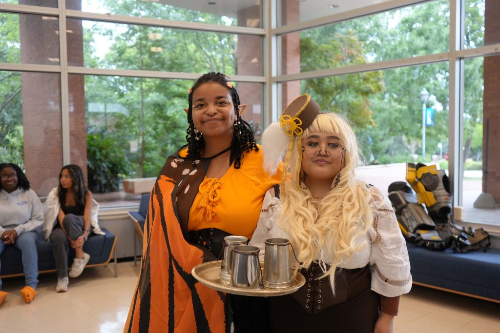 Two female college students stand in a large atrium dressed up in costumes.