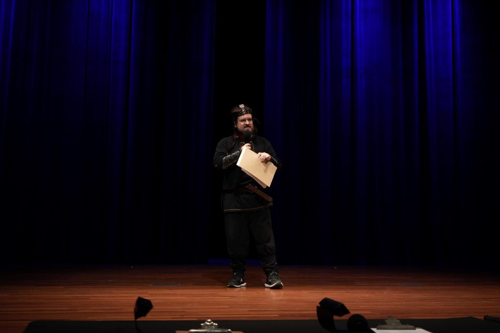 A man in a costume stands on a stage and talks to a large crowd.