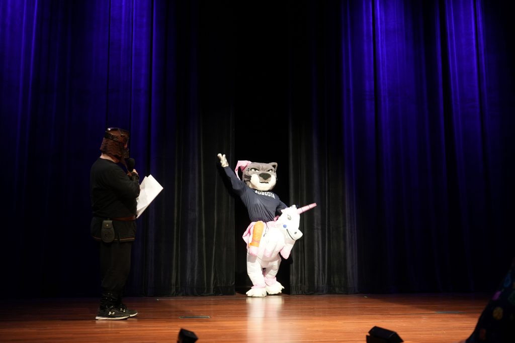 Maskot Universitas Augusta, Augustus, mengenakan kostum unicorn, berdiri di atas panggung. 