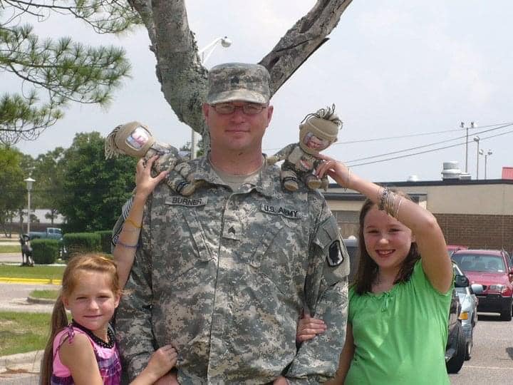 A man in an Army uniform stands in front of a tree with two young girls on either side of him holding stuffed soldiers with his face on them on his shoulders.