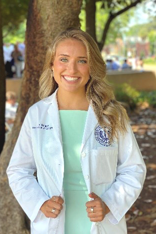 A woman in a light green dress and white coat smiles for the camera in front of a tree.