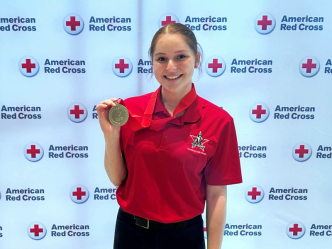 A female college student stands in front of a backdrop for the American Red Cross and holds up a medal.