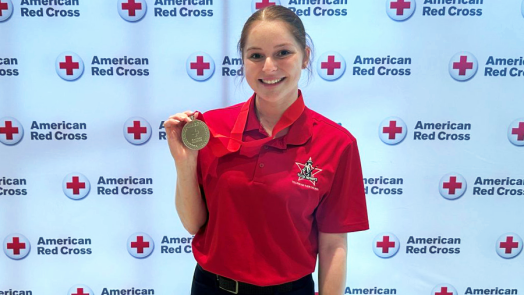 A female college student stands in front of a backdrop for the American Red Cross and holds up a medal.