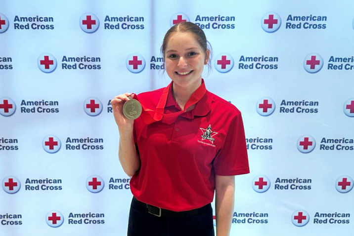 A female college student stands in front of a backdrop for the American Red Cross and holds up a medal.