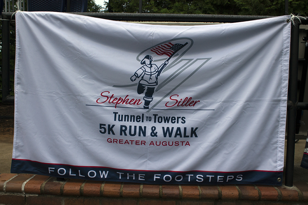 Picture of a white banner that says "Stephen Siller (in red) Tunnel to Towers 5K Run & Walk (in navy blue) Greater Augusta (in red)" in the center with a silhouette of a firefighter carrying an American flag. Across the bottom of the banner in white lettering against a navy background are the words "Follow the footsteps."