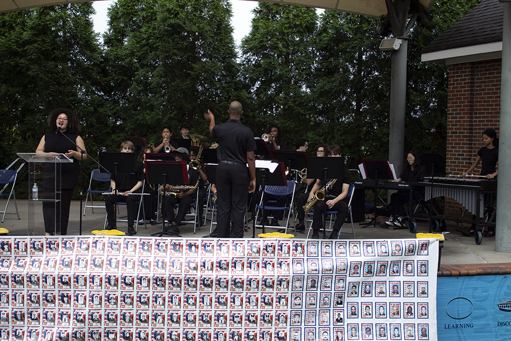 Woman sings on an outdoor stage next to a student band and its conductor. In front of the stage are photos of all of the firefighters killed during the 9/11 attacks.