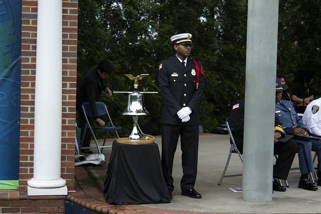 Man in full military regalia stands next to a silver bell with a gold eagle on top on a table