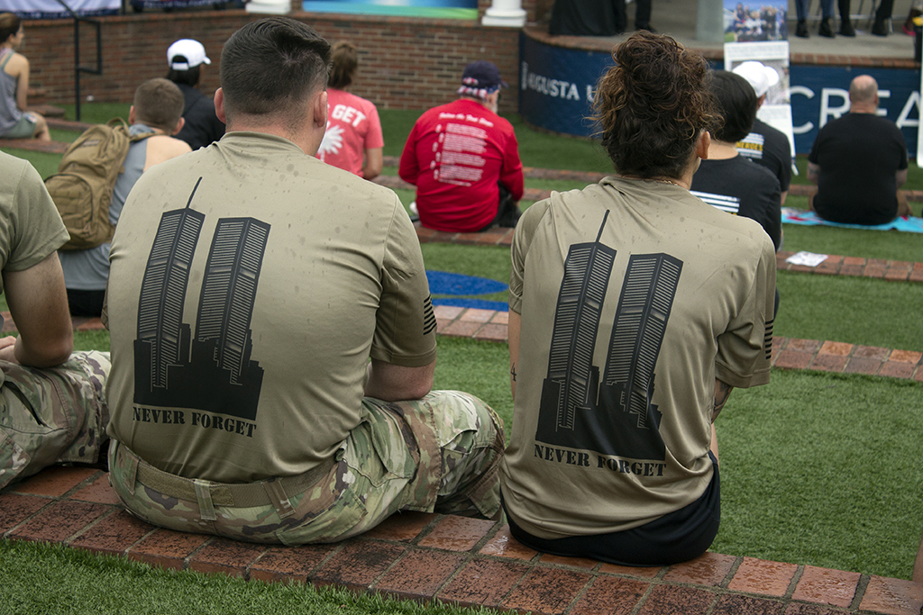 A man and woman sit in the rain wearing tan shirts depicting the Twin Towers and the words "Never Forget" underneath.