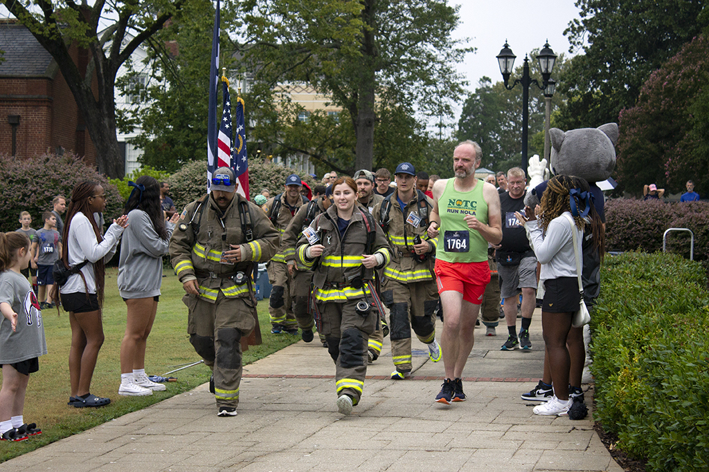 Firefighters in full firefighting attire begin to run the race next to a man in a tank top and shorts as people line the sidewalk cheering them on.