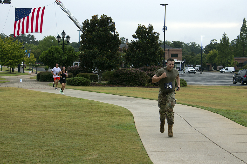 A man in a camo green shirt and camo pants runs down the sidewalk, while a man and woman in regular running attire try to catch up to him.