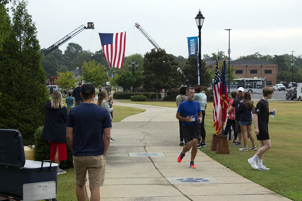 A man in a blue shirt, black shorts and orange running shoes crosses the finish line, as firetrucks in the background hold a large American flag over the path of the runners.