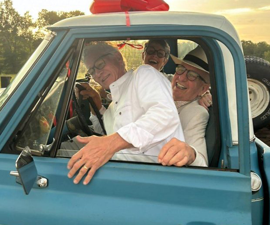 Three older adults laughing in the cab of a classic truck with a ribbon on top.