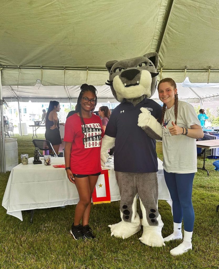 Two girls pose with a Jaguar mascot under a large tent.