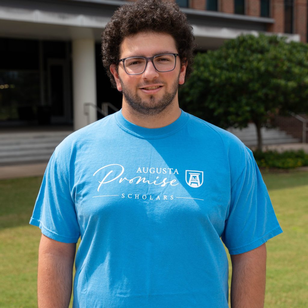 A male college student stands outside wearing t-shirt that says "Augusta Promise Scholars" across the chest.