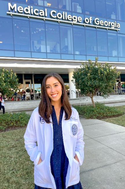 A woman in a blue dress and white coat stands in front of the Medical College of Georgia building.