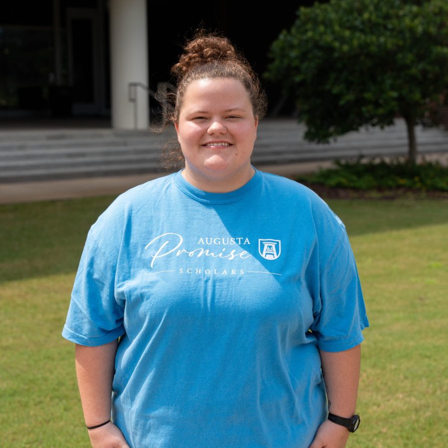 A female college student stands outside wearing t-shirt that says "Augusta Promise Scholars" across the chest.