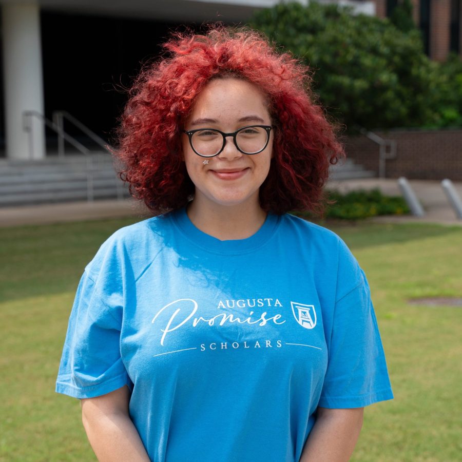 A female college student stands outside wearing t-shirt that says "Augusta Promise Scholars" across the chest.