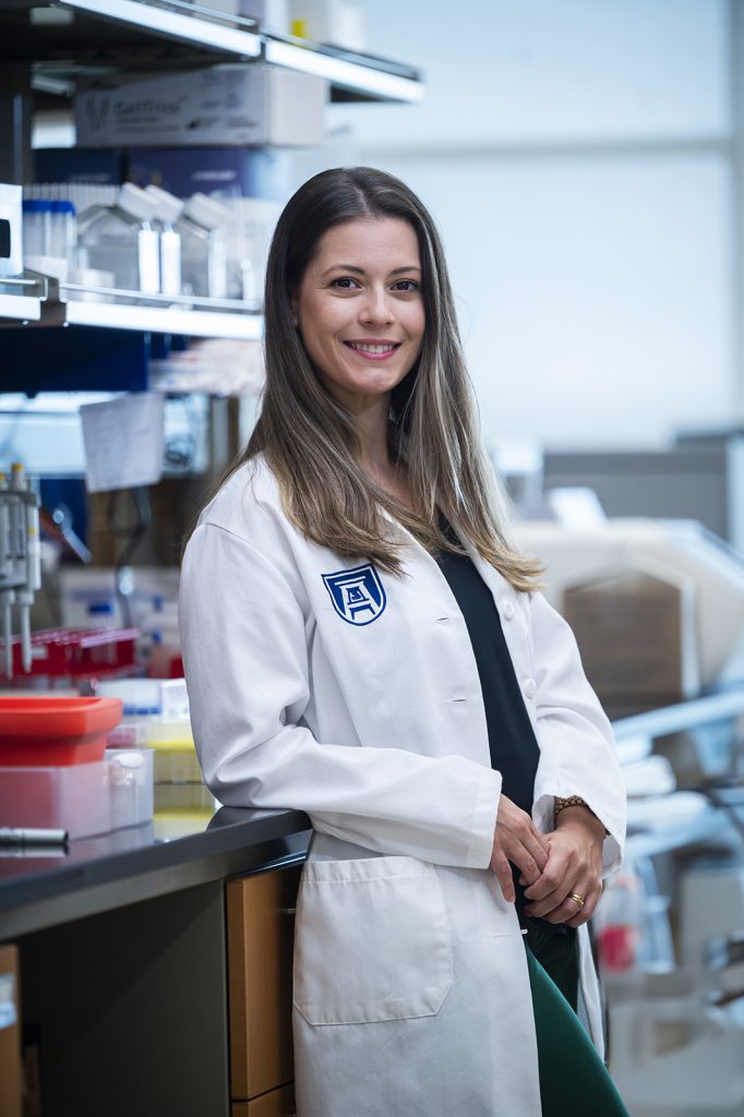 A woman wearing a white coat with a navy Augusta University bell tower logo leans against a counter in the lab and smiles at the camera.