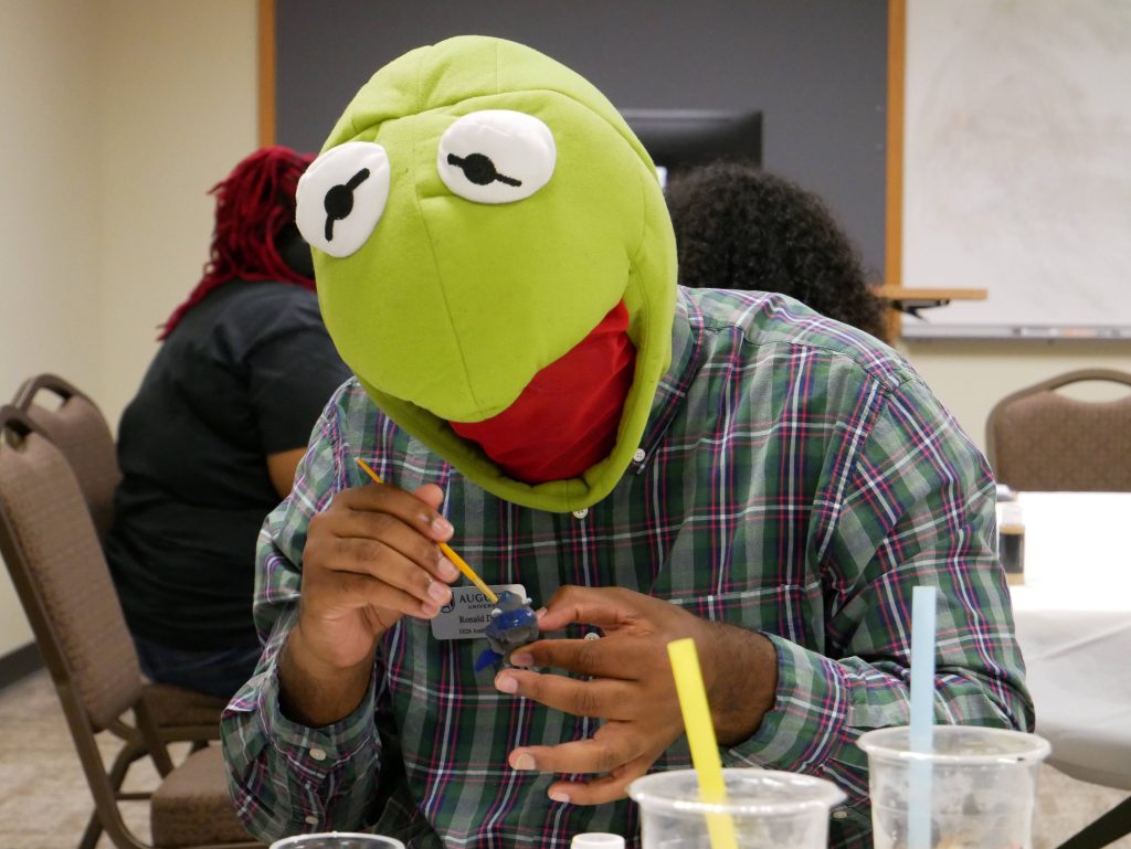 A male college student wearing a Kermit the Frog mask paints a mini figurine.