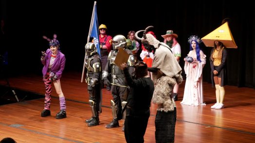 A large group of college students, all dressed in costumes from pop culture, stand on a stage during a costume contest.