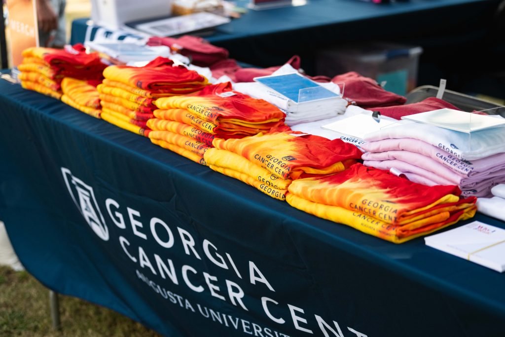 A table covered with a navy "Georgia Cancer Center Augusta University" tablecloth holds piles of pink or white or red/orange/yellow tie-dyed Georgia Cancer Center t-shirts.