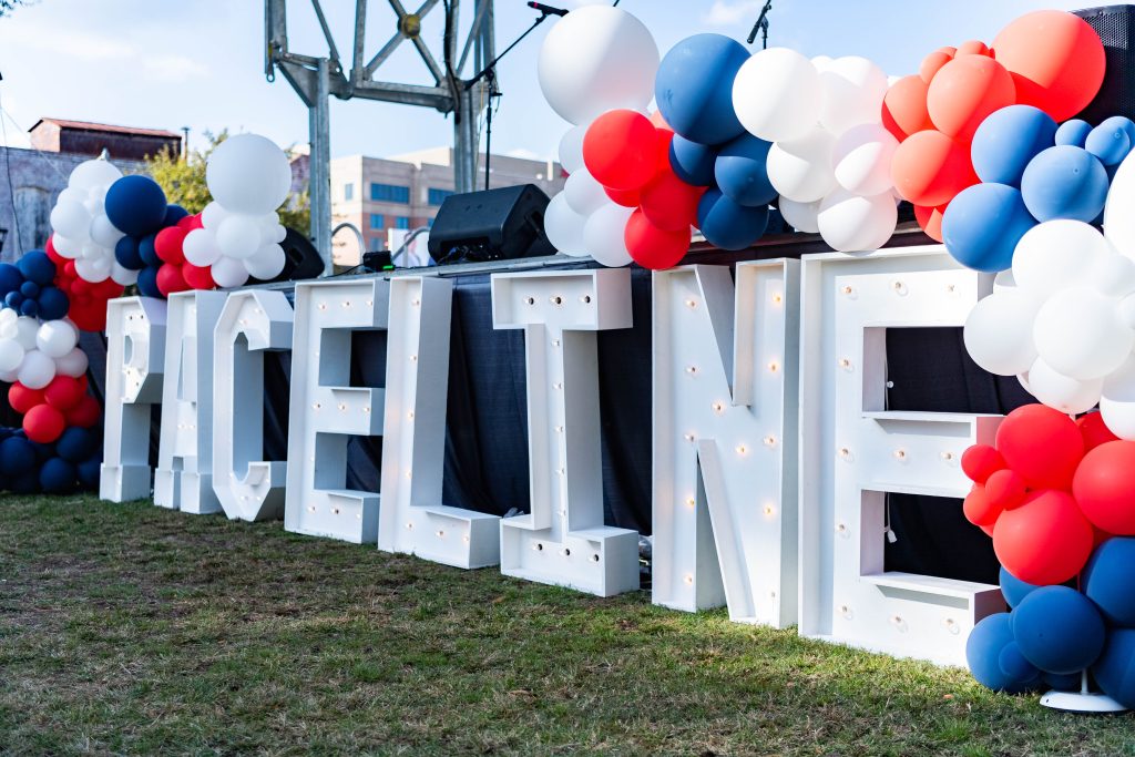 Giant white letters the height of a stage with lights in them spell out the word "Paceline" while clusters of white, red and blue balloons surround the letters.