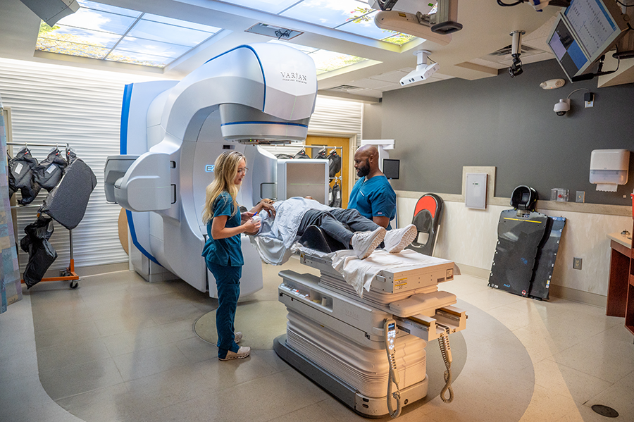 Radiation therapy students stand in a large room and use a scanning device that takes up most of the room to scan a patient.