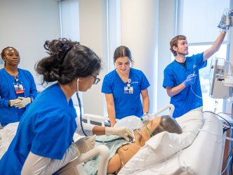 Four students in blue scrubs work on a dummy patient in the SIM center