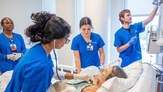 Four students in blue scrubs work on a dummy patient in the SIM center