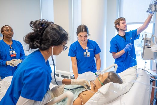 Four students in blue scrubs work on a dummy patient in the SIM center