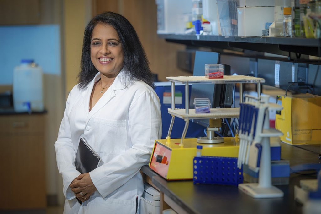 Woman in white coat holding notebook stands in lab and smiles