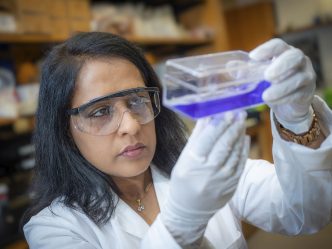 Woman wearing protective glasses and white gloves holds and stares at purple liquid