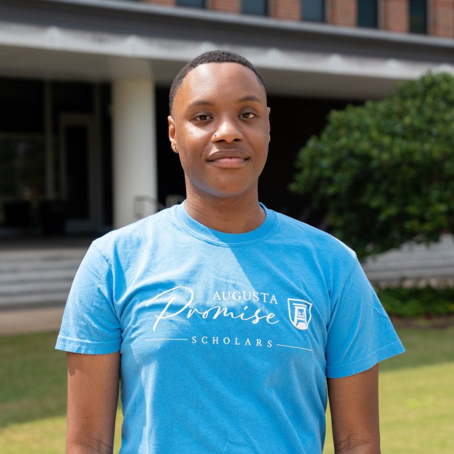 A male college student stands outside wearing t-shirt that says "Augusta Promise Scholars" across the chest.