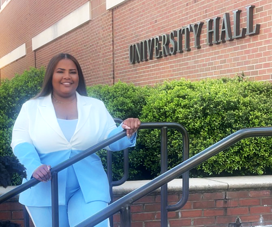 A woman in business attire stands outside along a railing. There is a sign for the name of the building behind her that reads "University Hall."