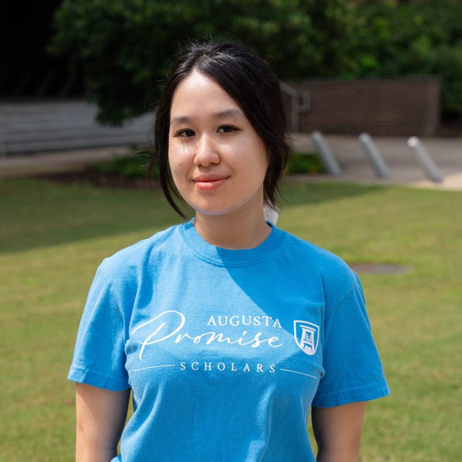 A female college student stands outside wearing t-shirt that says "Augusta Promise Scholars" across the chest.