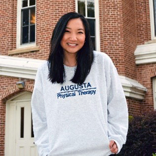 A woman wearing a light gray sweatshirt with the words "Augusta Physical Therapy" in blue lettering stands outside in front of a brick building.