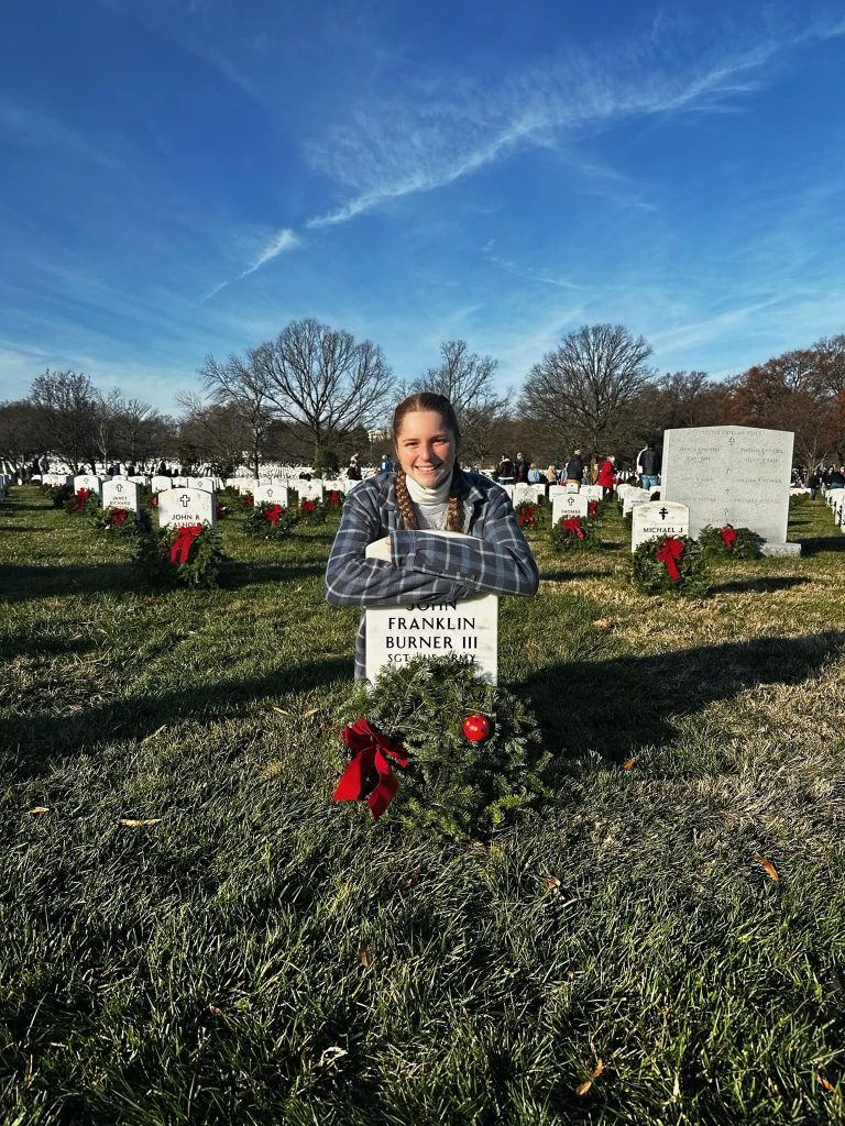 A woman leaned over a white headstone with a holiday wreath on the ground in front of it.