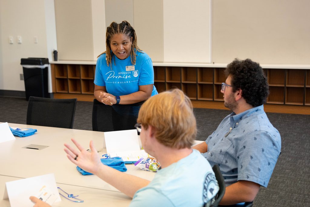 A woman leaning on a chair is talking with two male students.