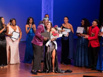 A group of male and female students dressed in evening wear is gathered around two other crowned students.