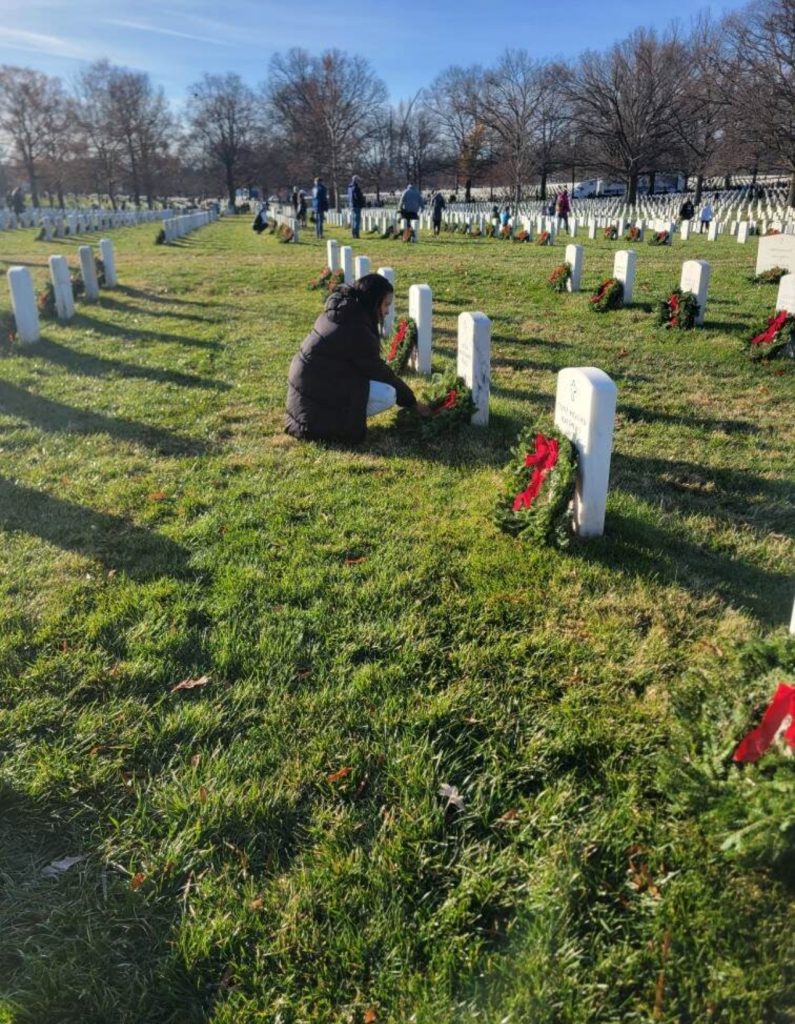 A woman is placing a wreath on the ground in front of a headstone.