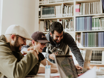 Three college-aged men gather around two laptops in a library.